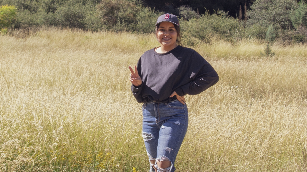 Yenifer Macias Lopez stands in a field smiling for the camera and making the peace hand sign